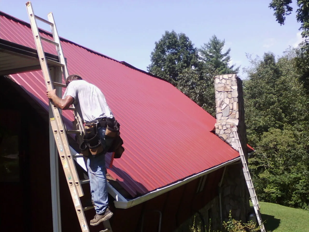 Roofer on a ladder beside a red metal roof and stone chimney — Soffit & Fascia Repair by Roofing Brothers in Gunnison